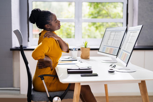 Woman at office desk reaching for upper back and shoulder in discomfort after prolonged sitting